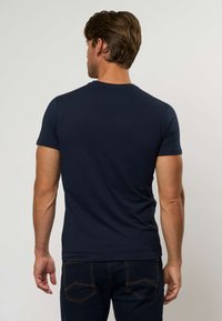 Tamsiai mėlynas trumpomis rankovėmis marškinėliai iš minkštos medžiagos, turintys apvalų kaklą ir prigludusį kirpimą. Dėvimi su tamsaus džinso kelnėmis.