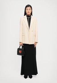 Woman wearing a cream blazer over a black long dress, holding a black handbag with a red heart design, standing against a plain white background.