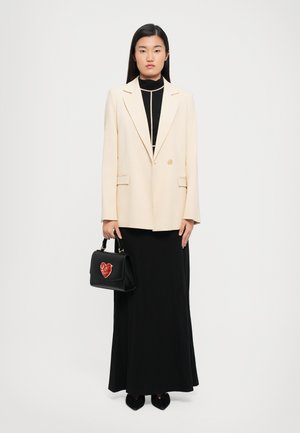 Woman wearing a cream blazer over a black long dress, holding a black handbag with a red heart design, standing against a plain white background.