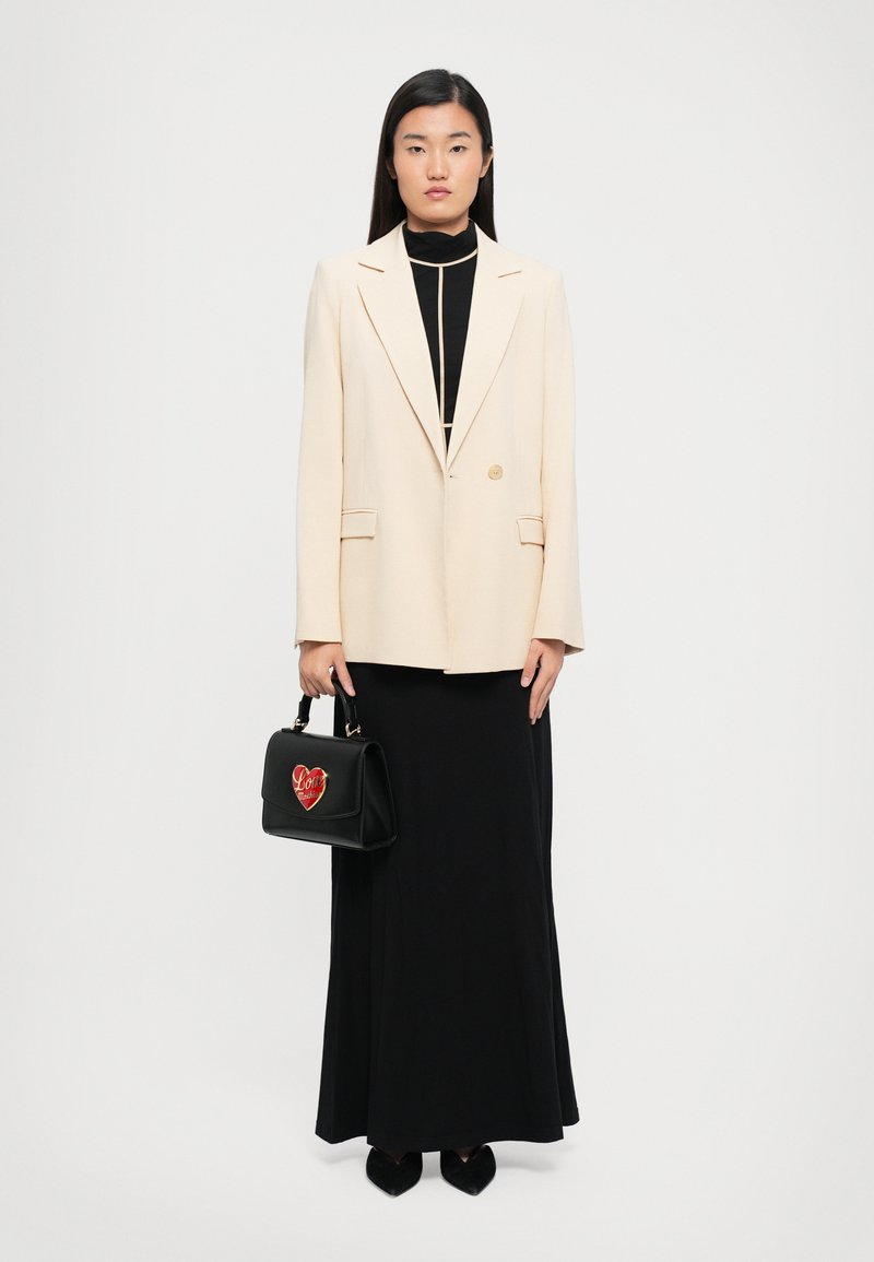 Woman wearing a cream blazer over a black long dress, holding a black handbag with a red heart design, standing against a plain white background.