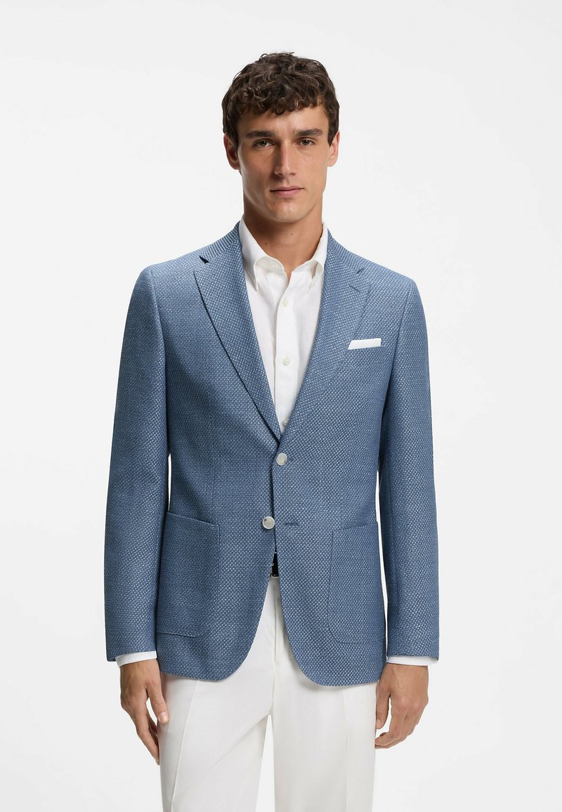 Homme portant un blazer bleu texturé, une chemise blanche et un pantalon blanc, debout devant un fond blanc uni.