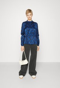Blusa blu con motivo a fantasia e vita peplum; abbinata a pantaloni grigi a gamba larga e una piccola borsa bianca. Le scarpe sono bianche con suola beige.