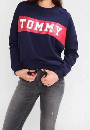 Mujer con sudadera azul marino con una franja diagonal roja y el texto blanco "TOMMY", combinada con vaqueros negros desteñidos. Manos cerca de la cintura.