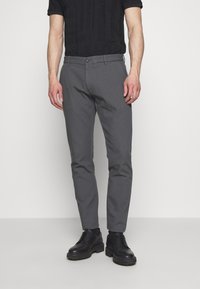 Grå smala byxor med flat front, tillverkade av ett mjukt tyg, med en enkel knappstängning och sidofickor. Svarta skor fulländar looken.