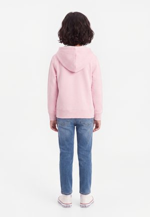 Barn med kort krøllet hår iført en pink hættetrøje, blå jeans og hvide sneakers står med ryggen til mod en hvid baggrund.