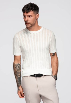 Homme aux cheveux courts et bouclés portant une chemise blanche en tricot à manches courtes et un pantalon beige, montrant des tatouages sur les deux avant-bras, regardant vers la gauche.