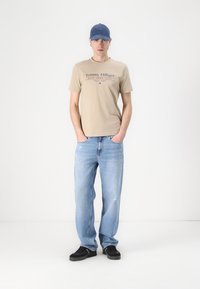 Lys beige bomull T-skjorte med "Tommy Hilfiger" logo, sammen med lyseblå denim jeans og sorte sneakers; person iført en blå caps.