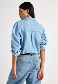 Lyseblå denimskjorte med op rullede ærmer, rygplissédetalje og spids krave, kombineret med højtaljede lyseblå jeans.