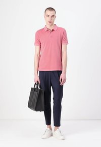 Roze polo, marineblauwe broek en witte sneakers. Model houdt een zwarte leren tas vast. Eenvoudig ontwerp, gladde textuur en nauwsluitende pasvorm.