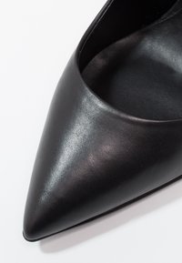 Gros plan sur une chaussure noire en cuir à bout pointu montrant une texture lisse et une doublure intérieure sur un fond blanc.