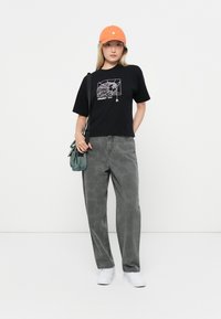 Black t-shirt with white spider web graphic, grey loose-fit pants, and an orange cap. White sneakers and a green shoulder bag complete the look.