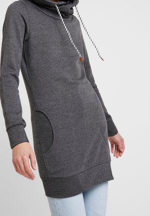 Person trägt einen langen, dunkelgrauen Kapuzenpullover mit weißen Kordeln und hellblauen Jeans, eine Hand an der Seite.