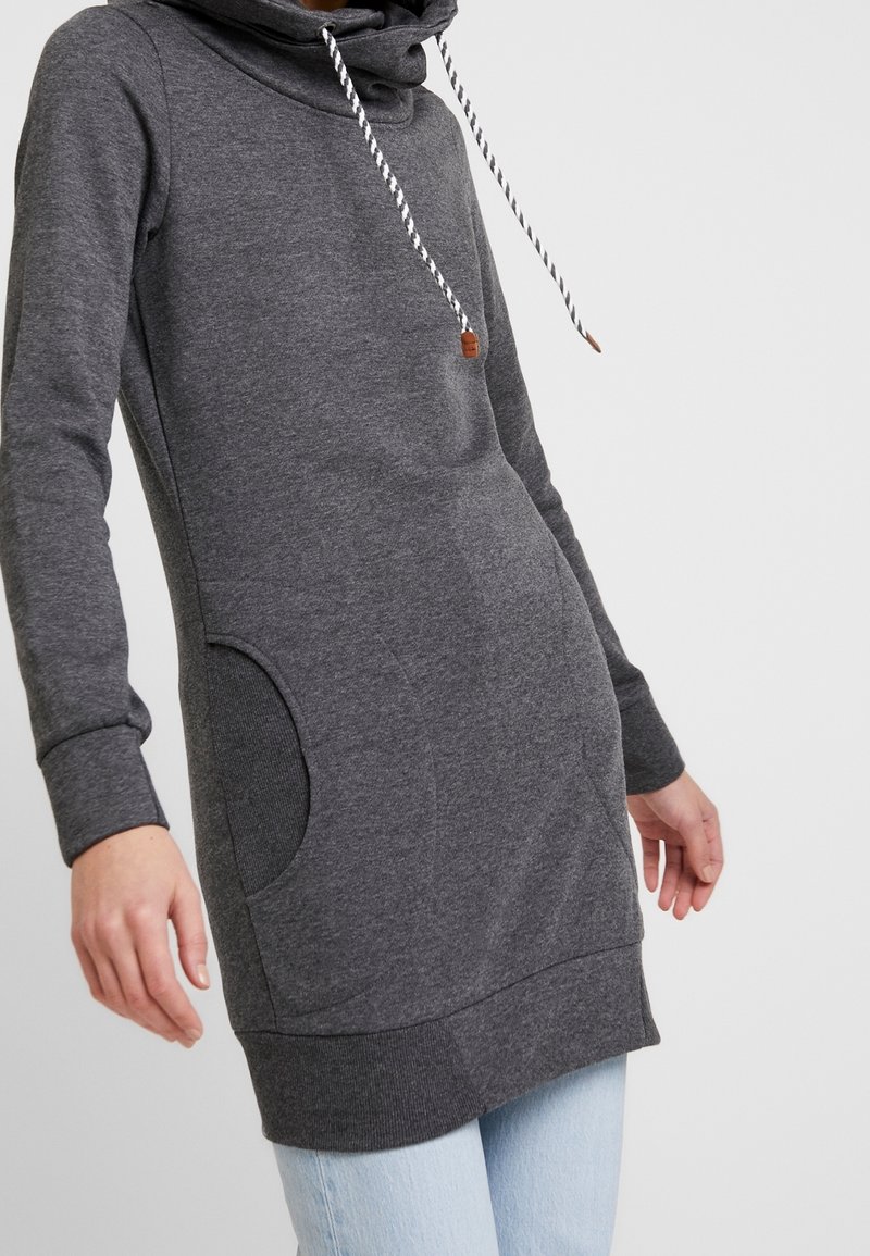 Personne portant un long sweat à capuche gris foncé avec cordons blancs et un jean bleu clair, une main le long du corps.