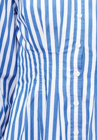 Blå og hvit skjorte med vertikale striper, knappelukking, lange ermer og innsvingt midje.