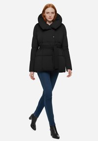 Giacca puffer nera imbottita con un grande cappuccio, vita cintata e chiusura a scatto frontale, abbinata a jeans skinny blu e stivaletti neri.