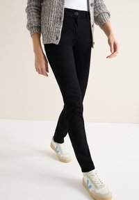 Jeans ajustados negros con cierre de botones, combinados con un cárdigan de punto beige y zapatillas blancas con detalles en azul.