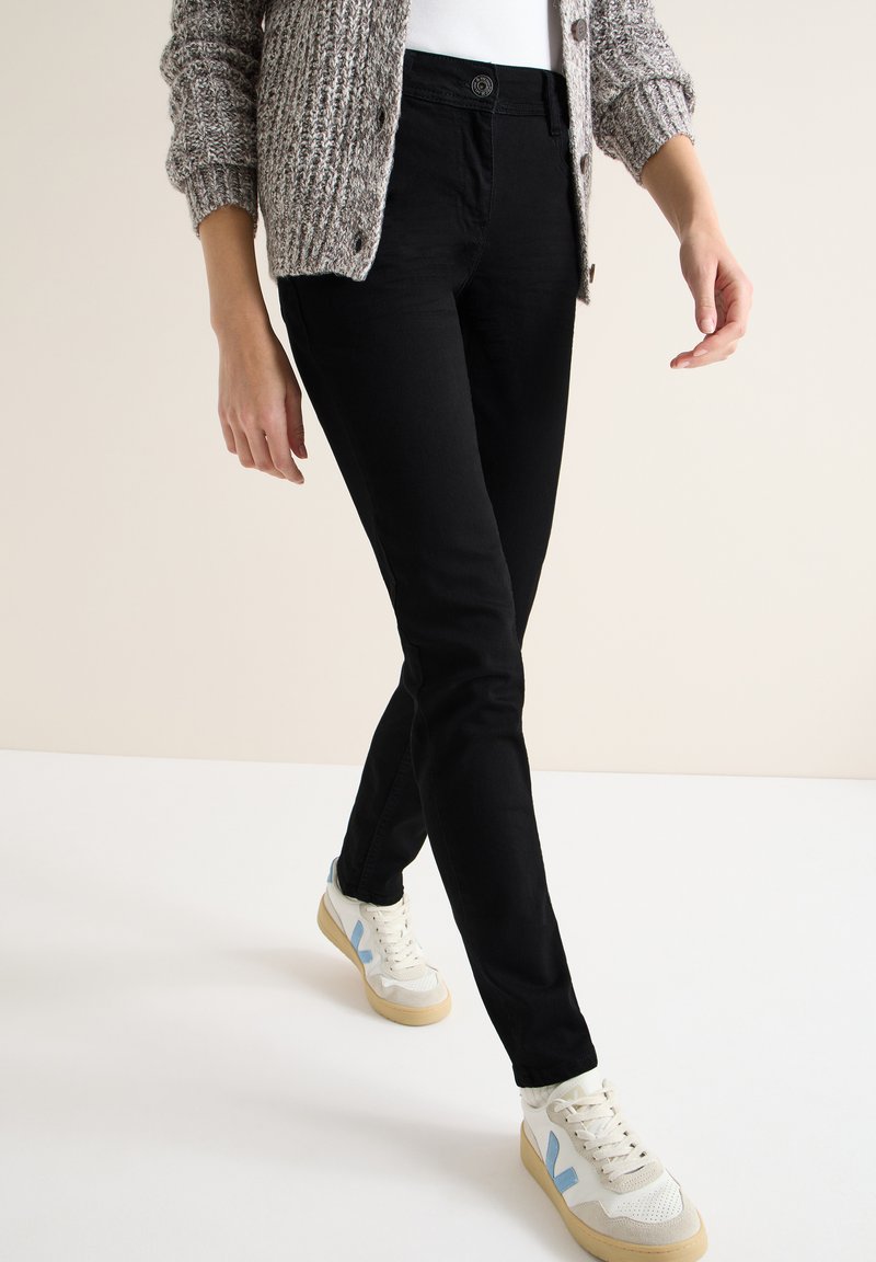 Jeans ajustados negros con cierre de botones, combinados con un cárdigan de punto beige y zapatillas blancas con detalles en azul.