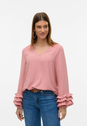 Blusa rosa chiaro con polka dot testurizzati, scollatura a V e maniche a volant a strati. Abbinata a jeans blu e una cintura marrone.