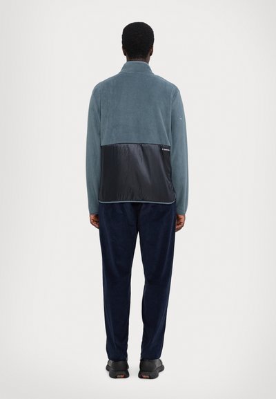 Veste en polaire avec un panneau en nylon noir en bas, de couleur bleu clair. Associée à un pantalon bleu foncé et des chaussures noires.