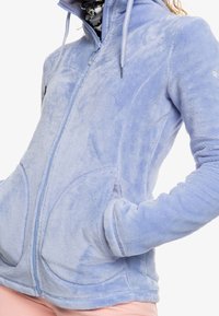 Vaaleansininen fleecetakki, jossa on täysi vetoketju, korkea kaulus, etutaskut ja tekstuurinen kangas, jossa on kaareva saumanmuotoilu hihoissa.