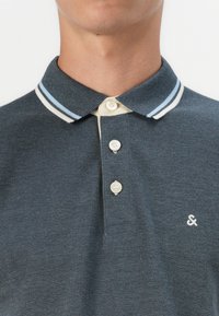 Polo de culoare albastru închis, dintr-un material texturat, având un guler bicolor cu dungi de culoare bleu deschis și crem, și trei nasturi. Logo mic pe piept.