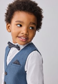 Conjunto formal para niños que incluye un chaleco texturizado azul, una camisa blanca y una pajarita con estampado. Los detalles notables incluyen botones marrones y un pañuelo de bolsillo.
