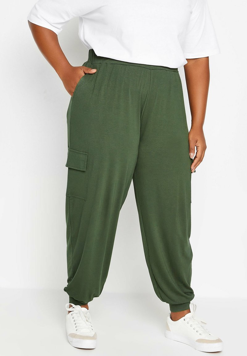 Personne de grande taille portant un pantalon de jogging cargo vert foncé et des baskets blanches, la main dans la poche, se tenant contre un fond blanc.