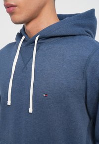 Μπλε φούτερ με λευκές κορδόνες, που διαθέτει ένα μικρό λογότυπο σε κόκκινο, άσπρο και μπλε. Μαλακό ύφασμα και χαλαρή εφαρμογή, με τσέπη καγκουρό.