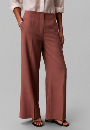 Donna che indossa pantaloni a vita alta e gamba larga color ruggine, camicia chiara a maniche lunghe arrotolate, sandali neri e braccialetto d’argento, in piedi con le mani nelle tasche.