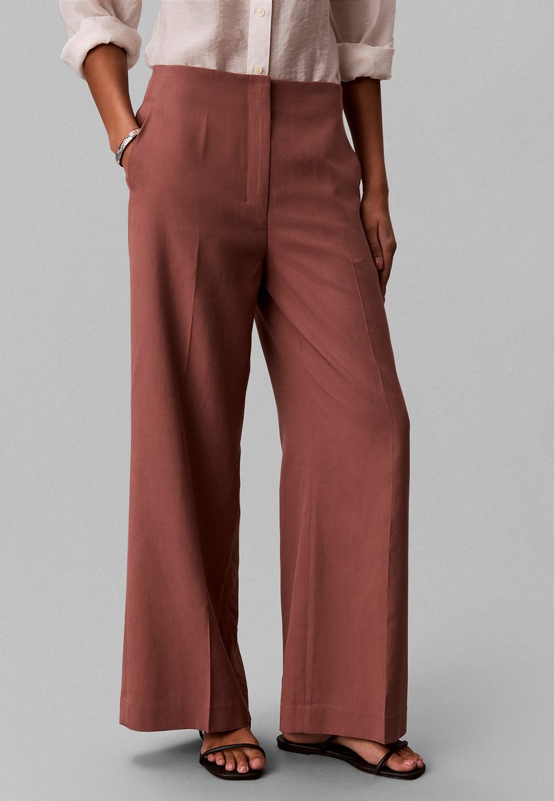 Femme portant un pantalon taille haute, jambes larges couleur rouille, chemise claire à manches longues retroussées, sandales noires et bracelet argenté, debout les mains dans les poches.
