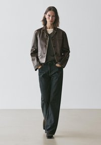 Brun læderjakke med en struktureret pasform, to forlommer og knapper. Bruges over en grå sweater og kombineres med mørke bredbenede jeans.