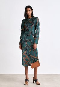 Grünes midi Kleid mit Kettenmuster, langen Ärmeln, Rundhalsausschnitt und lockerer Passform, kombiniert mit einer kleinen braunen Schultertasche und schwarzen High Heels.
