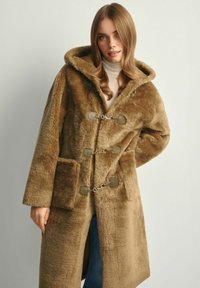 Brązowy płaszcz z sztucznego futra z kapturem, z metalowymi zapięciami w kolorze srebrnym, kieszeniami po bokach oraz miękką teksturą. Prosty, prostokątny fason.