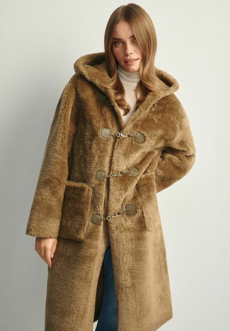 Brązowy płaszcz z sztucznego futra z kapturem, z metalowymi zapięciami w kolorze srebrnym, kieszeniami po bokach oraz miękką teksturą. Prosty, prostokątny fason.