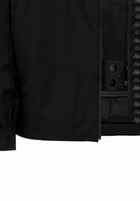 Veste noire partiellement ouverte montrant la doublure intérieure avec des boutons-pression marqués et un bord à motif logo sur le rabat de la fermeture éclair.