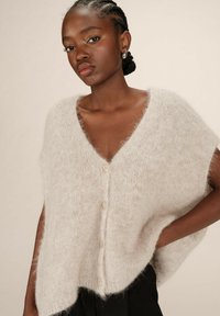 Cardigan en tricot beige clair avec un col en V, des manches courtes et cinq boutons sur le devant. Texture douce et duveteuse avec une coupe ample.