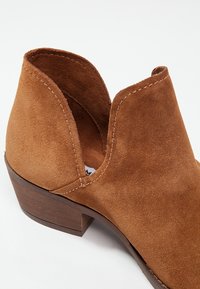 Bottine en daim marron avec un petit talon en bois empilé et découpes latérales ouvertes, montrant une texture lisse et une couture détaillée.