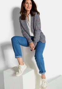 Femme assise sur un cube blanc, portant un cardigan en tricot gris, une blouse blanche, un jean bleu retroussé et des baskets blanches, regardant sur le côté.