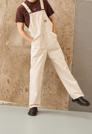 Person trägt cremefarbene Overalls und schwarze Stiefel, steht mit den Händen in den Taschen und einem angehobenen Bein an einer strukturierten Wand.