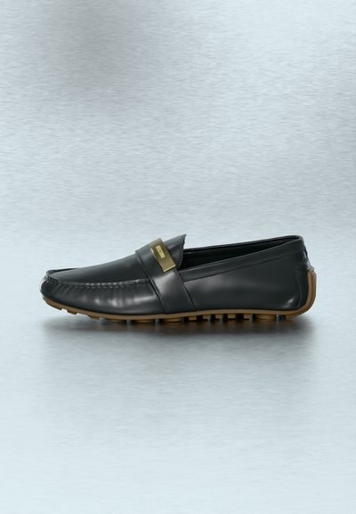 Mocassin en cuir noir avec semelle en caoutchouc marron et plaque en métal doré avec logo sur la bride, présenté sur une surface grise texturée.