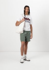 White t-shirt with red and blue text, olive green shorts, beige tote bag, white socks, and gray-and-white sneakers. Plain background.