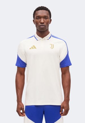 Homme portant un polo blanc et bleu de la Juventus avec le logo doré Adidas et l’emblème de l’équipe, debout devant un fond clair uni.