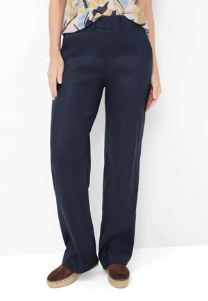 Personne portant un pantalon large bleu marine, des chaussures espadrilles marron, et un chemisier à motifs, debout devant un fond blanc.