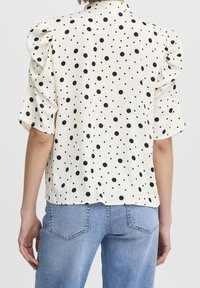 Short-sleeve cream blouse with black polka dots, featuring puffed shoulders and a relaxed fit, paired with light blue jeans.
