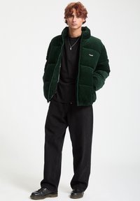 Green corduroy puffer jacket with high collar and zip closure, worn over a black t-shirt, paired with loose black pants and black shoes.