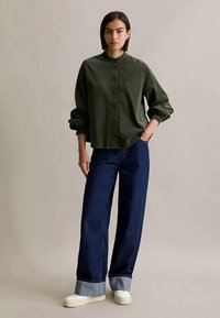 Mulher de pé com a mão no bolso, vestindo uma camisa verde escura com botões, calças de ganga largas e com punhos em azul escuro, e sapatos brancos, contra um fundo simples.