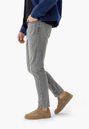 Man die een grijze slim-fit jeans, een marineblauwe jas en beige sneakers draagt, staand met één voet iets omhoog op een witte achtergrond.