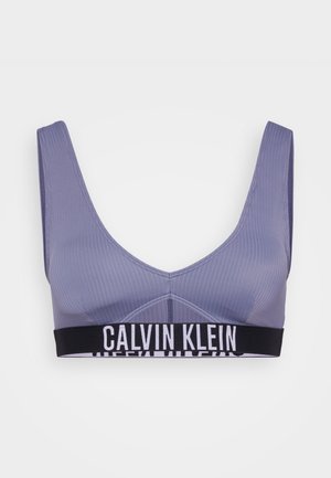 Calvin Klein sporta krūšturis gaisā purpursarkanā krāsā, ribota tekstūra, ar platām siksnām un melnu jostu, uz kuras redzams balts logo teksts.