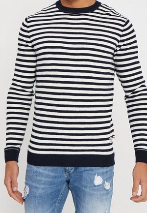 Mand iført en langærmet marineblå og hvid trøje med horisontale striber og slidte blå jeans, stående med armene afslappet ned langs siderne.