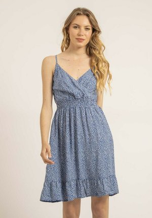 Jeune femme aux longs cheveux blonds ondulés portant une robe bleue sans manches à motif floral blanc, debout devant un fond uni.